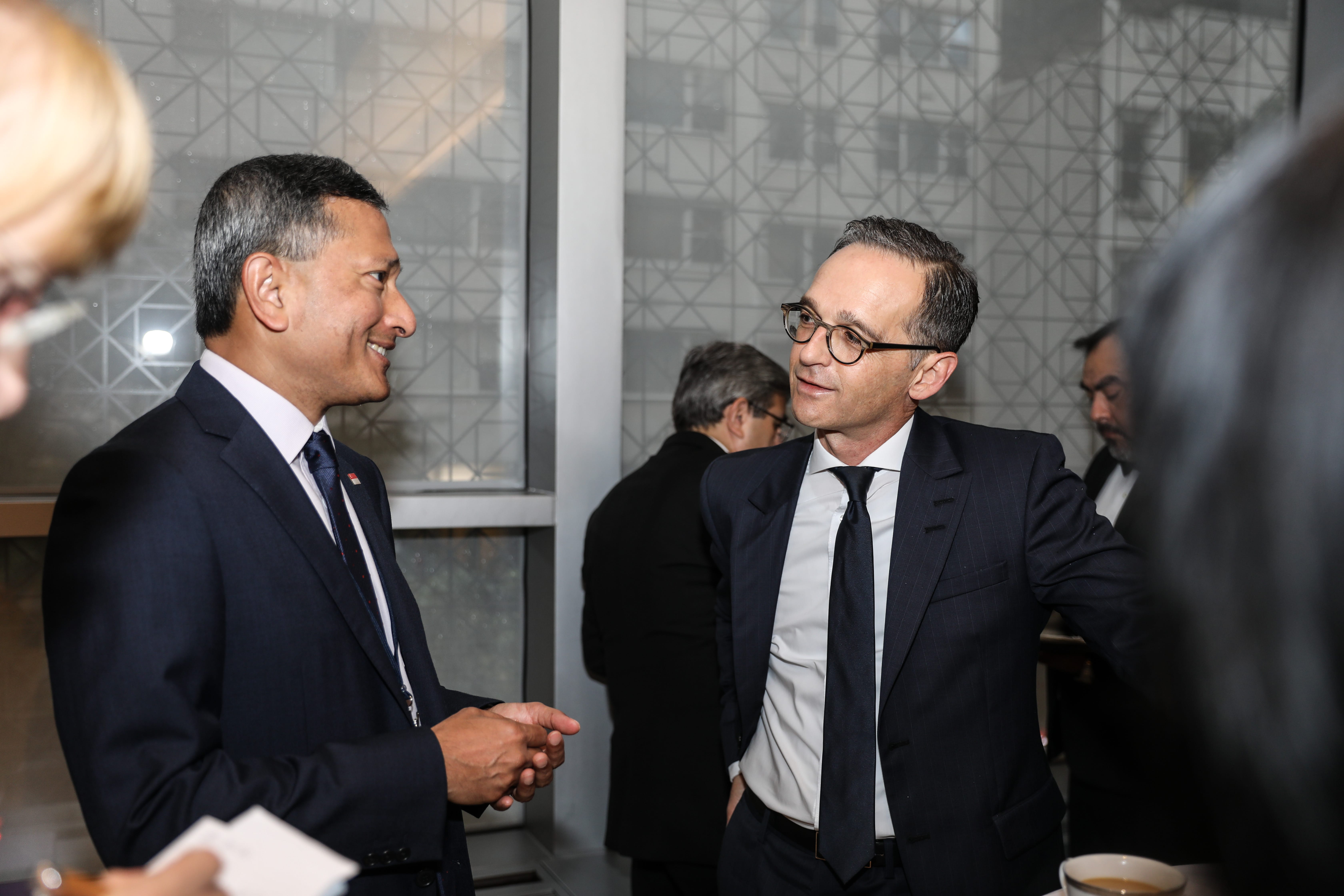 Two men in suits facing each other, having a conversation in front of a window.